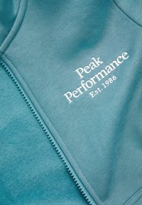 Teal sweatvest met ritssluiting, met geborduurd "Peak Performance" logo in het wit. Zachte textuur met een gladde stofafwerking.
