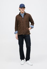 Jachetă maro cu fermoar purtată peste o cămașă în carouri albastre, combinată cu blugi închiși și adidași albi. Outfit-ul include o șapcă bleumarin cu logo.