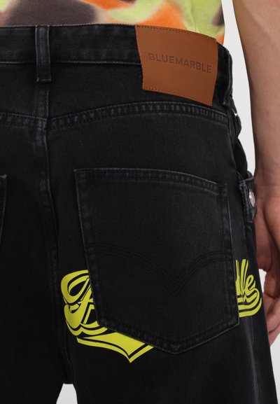 Zwarte spijkerbroek van denim met een grote gele print op de achterzak en een bruine leren patch met het label "BLUEMARBLE" bovenaan de tailleband.