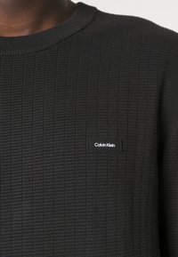 Maglione nero con texture, design a coste e collo rotondo, caratterizzato da una piccola etichetta nera Calvin Klein sulla parte anteriore.
