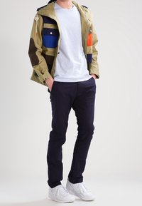 Kurtka wiatrówka w mieszanych kolorach, w tym oliwkowa zieleń, niebieski, pomarańczowy i granatowy. W zestawie z szarą koszulką i ciemnymi spodniami; białe trampki.