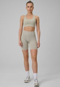 Jeune femme debout portant un soutien-gorge de sport beige, un short cycliste taille haute assorti, des chaussettes blanches et des baskets grises sur fond gris.