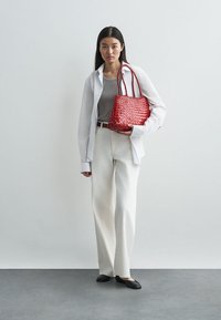 Piros szőtt kézitáska két fogantyúval, textúrázott felülettel. A modellen világosszürke felső, fehér, bő szabású nadrág és fekete lapos cipők vannak, semleges háttér előtt.