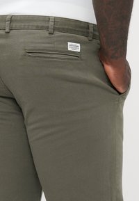 Pantalones verde oliva hechos de una tela suave con un corte ajustado. Presentan un bolsillo trasero y una pequeña etiqueta de marca cerca de la cinturilla.
