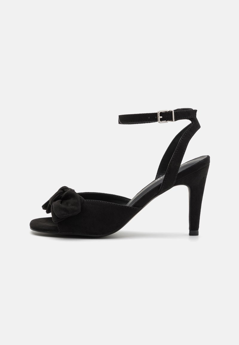 Sandale noire en daim à talon haut avec bride à la cheville, bout ouvert et nœud décoratif à l’avant, présentée sur fond blanc.
