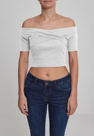 Frau trägt ein weißes schulterfreies, bauchfreies Oberteil und hoch taillierte dunkelblaue Jeans, steht vor einem schlichten grauen Hintergrund.