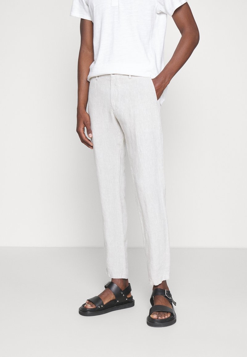 Homme portant un pantalon en lin blanc, une chemise blanche à manches courtes et des sandales en cuir noires, debout devant un arrière-plan uni.