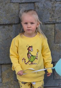 Gul sweatshirt med en tecknad kattdesign på framsidan. Barnet bär mönstrade gula byxor och håller en ljusblå ballong med band.