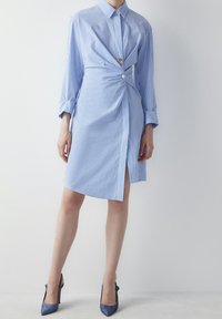 Abito camicia a righe blu chiaro e bianche con collo, dettaglio frontale torcigliato, accentuato da un unico bottone, lunghezza al ginocchio e maniche lunghe.