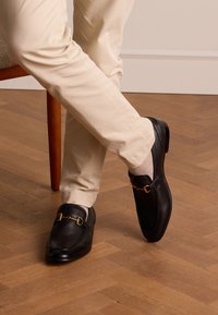 Black leather loafers with gold-tone hardware and a textured finish, paired with light beige trousers. Wood floor background.