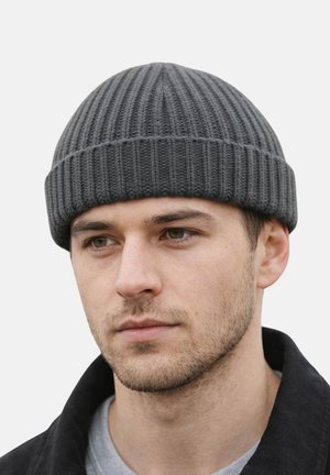 Junger Mann mit leichtem Stoppelbart, der eine graue, gerippte Strickmütze und eine schwarze Jacke trägt, blickt leicht zur Seite vor einem einfarbigen Hintergrund.