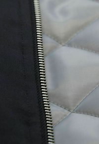 Schwarzer Stoff mit einem silbernen metallischen Reißverschluss; graue gepolsterte Fütterung sichtbar mit einer glatten Textur. Nahaufnahme des Reißverschlusses und des Stoffes.