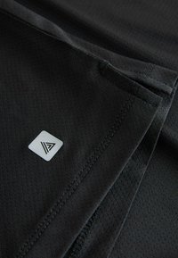 Tissu noir texturé avec un petit logo carré réfléchissant affichant une lettre "A" stylisée près des coutures cousues.