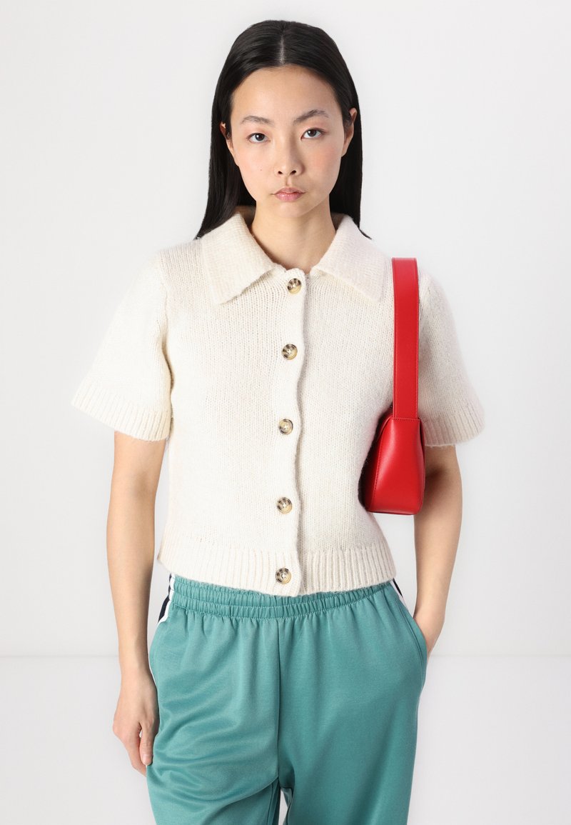 Kurzärmlige, cremefarbene Strickjacke mit einem kragenförmigen Ausschnitt, vorderen Knöpfen und geripptem Saum; kombiniert mit türkisfarbenen Jogginghosen und einer roten Handtasche.