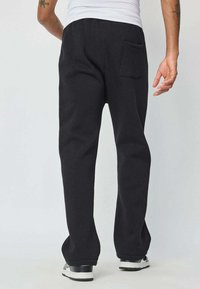 Pantalons de survêtement noirs en tissu doux, avec une coupe décontractée, une taille élastique et une poche arrière. Portés avec des baskets.