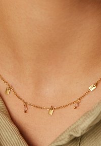 Gouden halsketting met kleine rechthoeken en kralen, met een delicate kettingontwerp, rustend tegen de huid met een gestructureerde stoffen achtergrond.