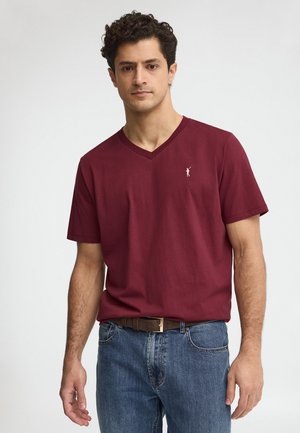 Hombre con cabello corto y rizado, vestido con una camiseta de cuello en V marrón y jeans azules, sobre un fondo blanco liso.