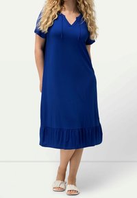 Blauwe katoenen jurk met een V-hals en korte mouwen, met een rucheszoom. Draag kennis met lichtgekleurde sandalen. Glad textuur.