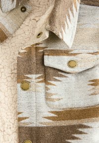 Close-up van een bruin en beige gestreepte wollen jas met metalen drukknoopsluitingen en een crèmekleurige sherpa voering.