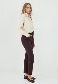 Camicia crema con bottoni, pantaloni aderenti marrone scuro e ballerine nere perforate. L'outfit presenta un design sagomato e texture morbide.