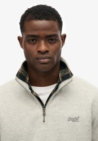 Szary polar z suwakami, z wysokim kołnierzem, noszony na koszuli w kratę. Posiada szczegóły w szyciu i logo na klatce piersiowej.