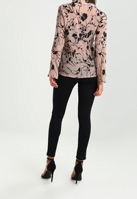 Blusa rosa cipria con ricami floreali neri, maniche lunghe e colletto, abbinata a jeans neri aderenti e scarpe con tacco alto.