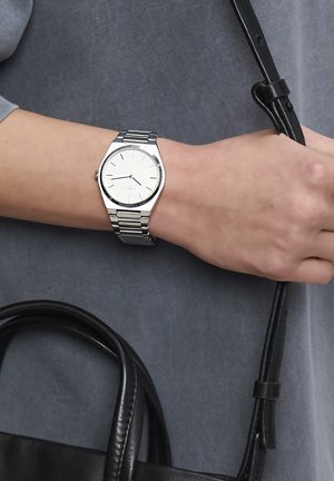Silberner Metalluhren mit rundem Zifferblatt, weißem Ziffernblatt, minimalistischen schwarzen Markierungen und einem Gliederarmband. Getragen an einer Hand, die eine schwarze Tasche hält.
