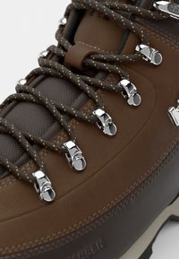 Bota de senderismo de cuero marrón con herrajes metálicos, texturas en contraste de marrón oscuro y claro, y cordones marrones con acentos amarillos.