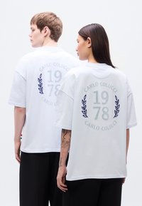 Λευκά βαμβακερά μπλουζάκια σε μεγάλο μέγεθος, με το κείμενο «CARLO COLUCCI 1978» και σχέδιο δάφνης στο πίσω μέρος. Χαλαρή εφαρμογή με κοντά μανίκια και απαλό βαμβακερό υλικό.