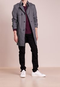 Szary trencz z przodu zapinany na guziki, z bocznymi kieszeniami i mankietami na guziki, noszony na bordowej bluzie. Do tego czarne jeansy i białe trampki.