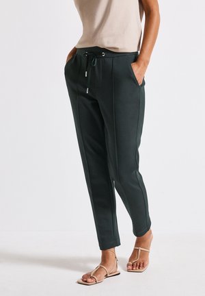 Woman wearing dark green tapered pants with front pockets and beige strappy sandals, standing with hands in pockets.