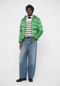 Zöld pufi dzseki, cipzárral, csíkos pulóver felett viselve. A laza szabású kék farmernadrág és fekete cipő egészíti ki az öltözetet.