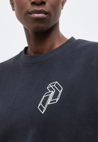 Κοντινό πλάνο ατόμου που φοράει σκούρο πουκάμισο με λευκό γεωμετρικό σχέδιο κύβου στο στήθος, μπροστά σε απλό φόντο.