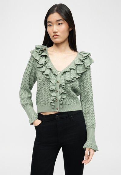 Vrouw die een saliegroene gebreide cardigan draagt met geruite afwerking en knopen, gecombineerd met een zwarte broek, staand met één hand in de zak.