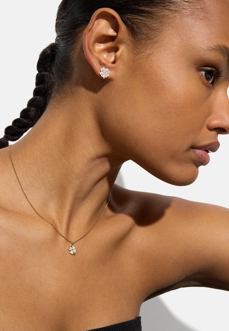 Złoty naszyjnik z małym, kwiatowym wisiorkiem; diamentowe akcenty. Małe kolczyki w kształcie kwiatu doskonale uzupełniają naszyjnik. Gładkie tło skóry.