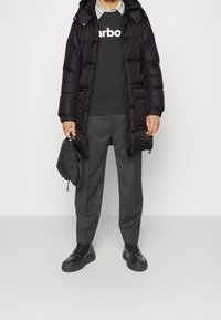 Svart puffjacka med huva, grå sweatshirt med logotyp, grå byxor och svarta skor. Håller en liten svart väska.