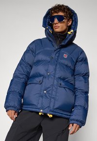 Navy blue puffer jacket with a hood, featuring yellow drawstrings, snap buttons, and two front pockets. Smooth, shiny texture.