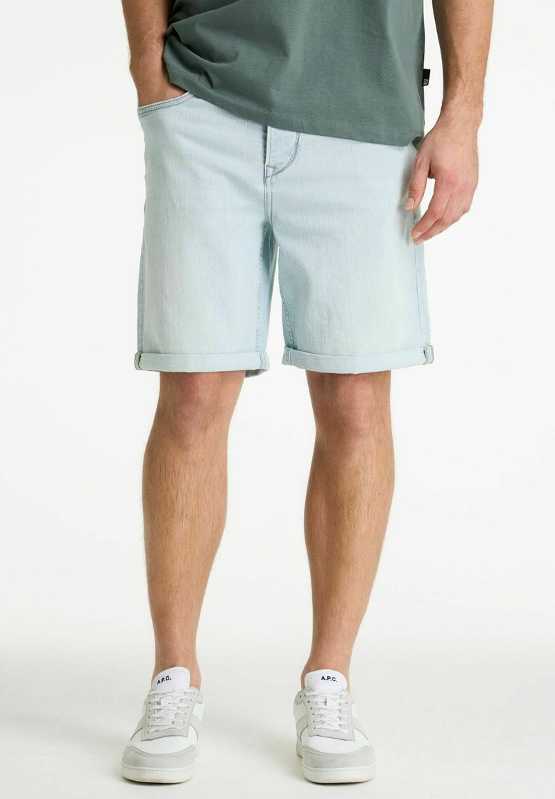 Man draagt lichtblauwe denimshorts met opgerolde zoom, groene T-shirt en witte sneakers, staat met één hand in zijn zak tegen een witte achtergrond.