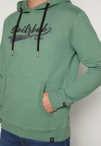 Grüne Kapuzenjacke aus weichem Stoff, mit einer vorderen Tasche, schwarzen Kordeln und einem grafischen Design mit "EST. 2017" in dunkler Schrift.