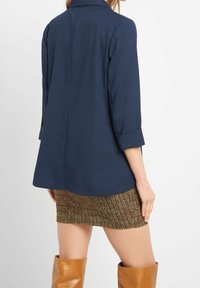 Lehký blejzr tmavě modré barvy s límečkem, s ohrnutými rukávy a rovný střihem, kombinovaný s vzorovanou sukní a hnědými kozačkami ke kolenům.