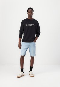 Sweatshirt preto com texto branco, calções de ganga azul claro, meias pretas e ténis bege com sola de borracha, posicionados contra um fundo branco.