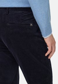 Granatowe spodnie kordrowe z pionowymi paskami, wyposażone w kieszeń z suwakiem z tyłu oraz subtelną metalową aplikacją z logo. Połączone z jasnoniebieskim swetrem.