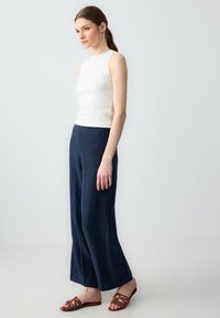 Witte geribbelde mouwloze top gecombineerd met losse, donkerblauwe linnen broeken en bruin leren sandalen met kruislings banden.