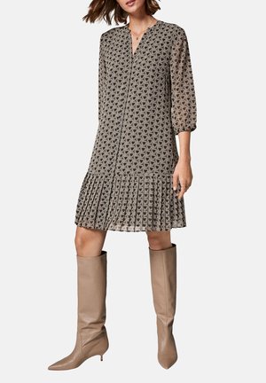 Femme portant une robe à motifs géométriques avec un ourlet plissé et des manches trois-quarts, associée à des bottes beige montant jusqu'aux genoux.