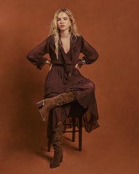 Lång, mörk burgundradräkt med volangkant, kombinerad med knähöga bruna stövlar prydda med nitar. Sitter på en träpall mot en brun bakgrund.