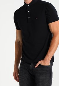 Man wearing a fitted black polo shirt with three white buttons and dark jeans, hand in pocket against a white background.
