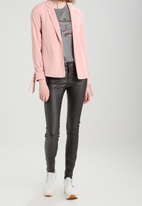 Blazer rosa claro con un corte relajado y detalles de lazo en las mangas, combinado con una camiseta gráfica gris y jeans ajustados negros. Zapatillas blancas.