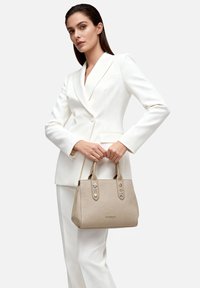 Donna con abito bianco su misura che tiene una borsa beige con dettagli metallici, in piedi contro uno sfondo chiaro semplice.