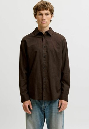 Jeune homme portant une chemise brun foncé à boutons et un jean bleu clair délavé, debout devant un fond clair uni.