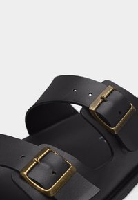 Μαύρο δερμάτινο σανδάλι με δύο ρυθμιζόμενους ιμάντες που διαθέτουν τετράγωνα μπρονζέ αγκράφες σε απλό φόντο.